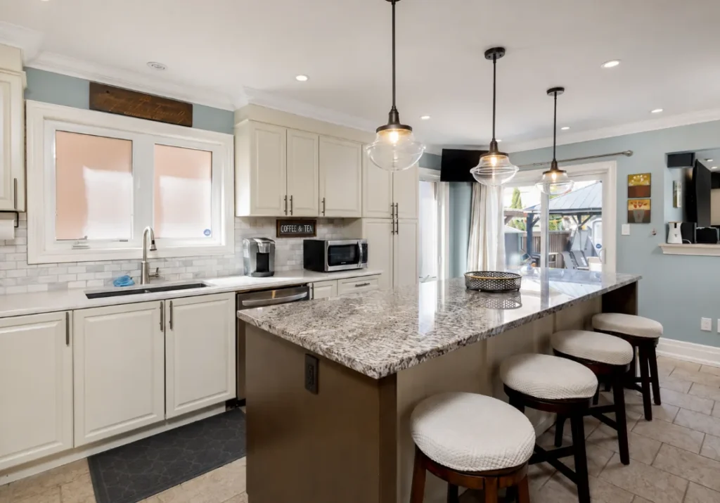 White kitchen with a stone-topped island, upholstered stools and glass pendant lights.